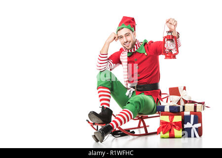 L'homme en costume de sorcière assis sur un traîneau près de tas de cadeaux et le holding Red Lantern isolated on white Banque D'Images