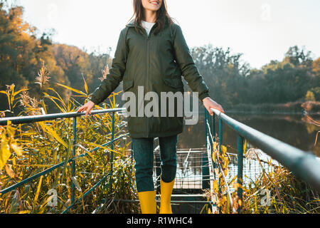 Vue partielle de la jeune femme en veste et bottes en caoutchouc jaune posant près de l'étang dans le parc Banque D'Images