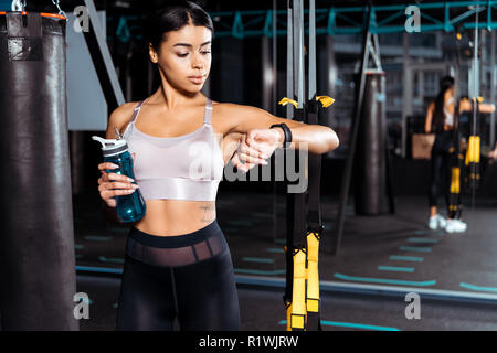 Sportive fantastique girl holding water bottle while looking at fitness tracker dans une salle de sport Banque D'Images