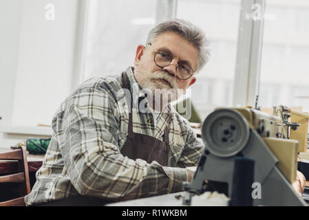 Portrait de femme mature en tailleur et lunettes tablier assis près de la machine à coudre au studio Banque D'Images