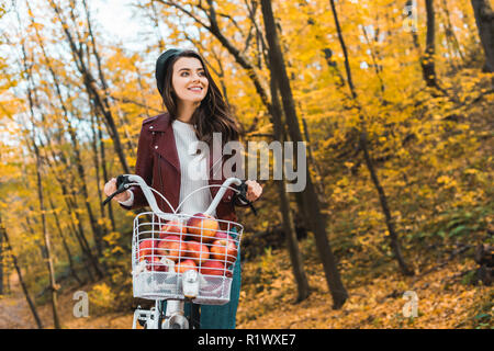 Heureux à la mode fille en blouson de cuir portant location avec panier rempli de pommes rouges dans le parc en automne Banque D'Images