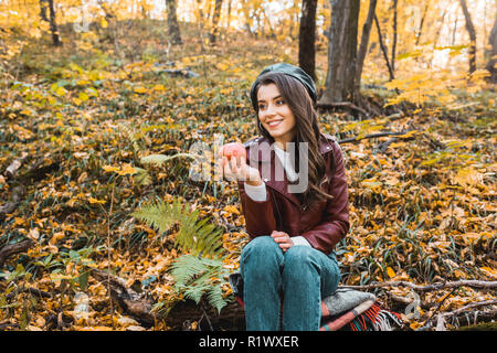 Smiling girl élégante en cuir veste assis sur la couverture et l'holding red apple in forest Banque D'Images