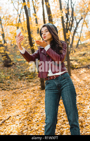 Low angle view of belle femme veste en cuir élégante en soufflant de l'air et baiser en tenant sur smartphone selfies en forêt Banque D'Images