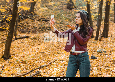 Jeune femme en soufflant de l'air veste en cuir élégant et kiss sur smartphone selfies en parc d'automne Banque D'Images