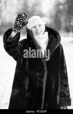 Un hiver portrait of a smiling senior femme portant un chapeau de laine, une écharpe et des gants de couleur, saluant avec sa main, avec un fond de neige Banque D'Images