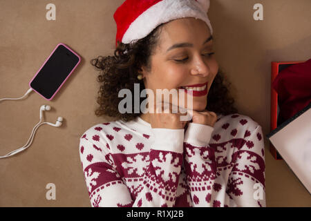 Smiling African American Woman wearing Santa hat and Christmas sweater couché et regardant don fort. Banque D'Images