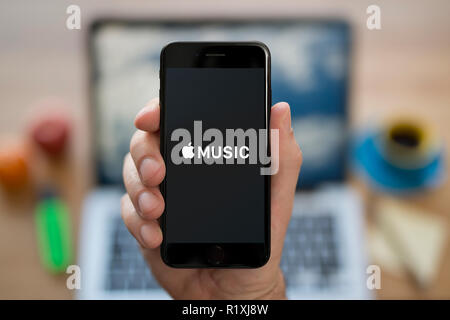 Un homme se penche sur son iPhone qui affiche le logo Apple Musique, tout en s'assit à son bureau informatique (usage éditorial uniquement). Banque D'Images