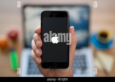 Un homme se penche sur son iPhone qui affiche le logo Apple, alors qu'assis à son bureau de l'ordinateur (usage éditorial uniquement). Banque D'Images