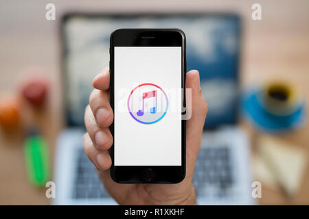 Un homme se penche sur son iPhone qui affiche le logo Apple iTunes, tout en s'assit à son bureau informatique (usage éditorial uniquement). Banque D'Images