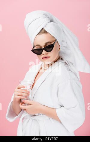 Jeune femme surdimensionnée en peignoir, lunettes et towel on head holding verre isolé sur Rose Banque D'Images