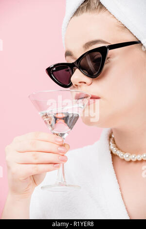 Close-up view of young woman in bathrobe surdimensionnée, lunettes de soleil et serviettes sur la tête de la consommation d'alcool et de verre isolé sur Rose Banque D'Images