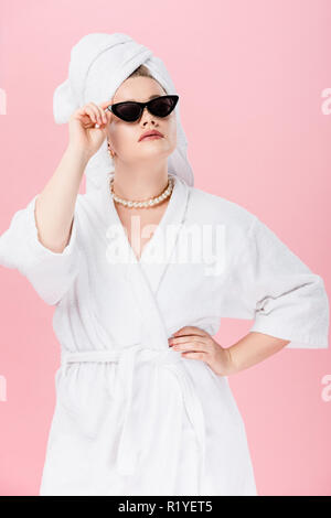 Jeune femme surdimensionnée en peignoir, lunettes et towel on head posing with hand on taille isolé sur pink Banque D'Images