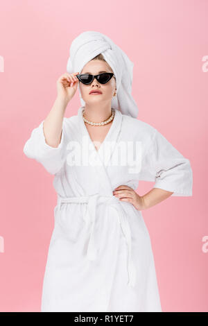 Jeune femme surdimensionnée en peignoir, lunettes et towel on head standing with hand on taille isolé sur pink Banque D'Images