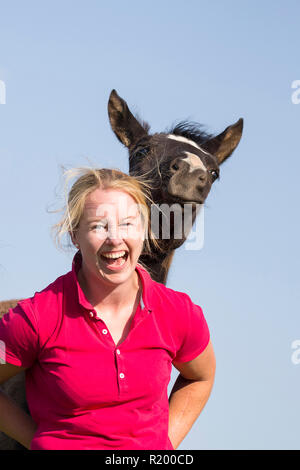 Warmblood. Laughing woman avec poulain ludique sur un pâturage. Allemagne Banque D'Images