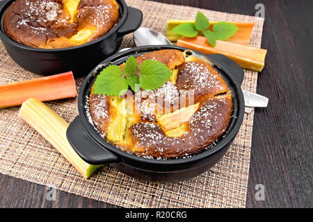 Klafouti tarte à la rhubarbe en noir brazier, menthe et rhubarbe tiges sur l'osier serviette sur planche en bois historique Banque D'Images