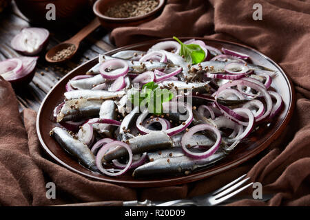 Anchois salé mariné à l'oignon rouge sur une plaque de faïence les graines de coriandre, brown tissu, fourchette et couteau sur une vieille table en bois rustique, vue Banque D'Images