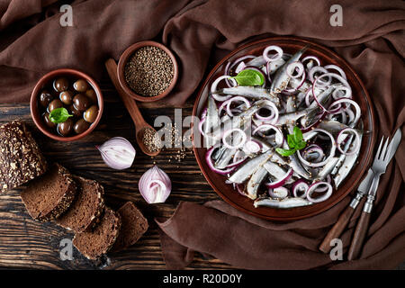 Anchois salé mariné à l'oignon rouge sur une plaque de faïence. Les graines de coriandre, brown tissu, fourchette et couteau sur une vieille table en bois rustique, vue Banque D'Images