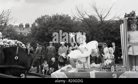 Années 1950, historique, une petite fille portant une couronne et robe et tenant une baguette étoile fée est assise sur une petite chaise sur un cheval dessiné flottent dans un carnaval, observé par les spectateurs sur le côté de la rue, England, UK. Banque D'Images