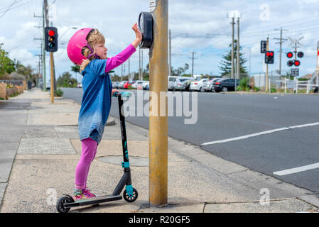 Jeune fille en costume rose et rose casque équitation un kick-scooter appuyant sur le bouton à pedestriam crossing Banque D'Images