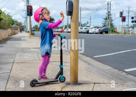 Jeune fille en costume rose et rose casque équitation un kick-scooter appuyant sur le bouton à pedestriam crossing Banque D'Images