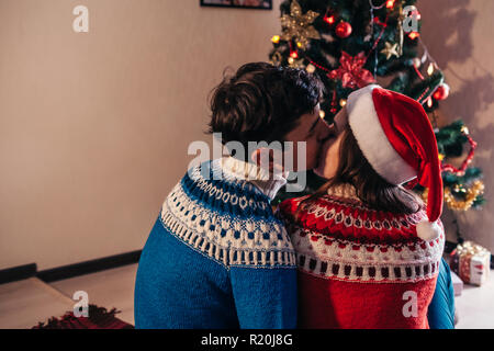 Couple amoureux assis près d'un arbre de Noël, wearing Santa's hat et s'étreindre. Les jeunes de célébrer le Nouvel an à la maison le soir Banque D'Images