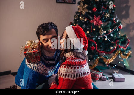 Couple amoureux assis près d'un arbre de Noël, wearing Santa's hat et s'étreindre. Les jeunes de célébrer le Nouvel an à la maison le soir Banque D'Images