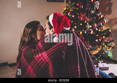 Couple amoureux assis près d'un arbre de Noël, wearing Santa's hat et s'étreindre. Les jeunes de célébrer le Nouvel an à la maison le soir Banque D'Images