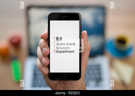 Un homme se penche sur son iPhone qui affiche le logo du Ministère de l'actuaire du gouvernement, tout en s'assit à son bureau informatique (usage éditorial uniquement). Banque D'Images