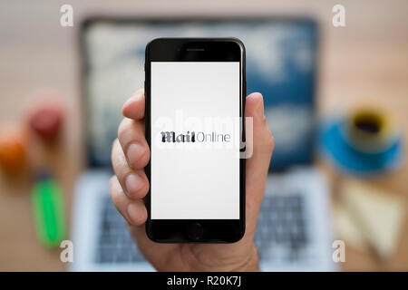 Un homme se penche sur son iPhone qui affiche le logo en ligne Courrier, tandis que s'assit à son bureau informatique (usage éditorial uniquement). Banque D'Images