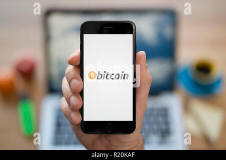 Un homme se penche sur son iPhone qui affiche le logo de Bitcoin, tandis qu'assis à son bureau de l'ordinateur (usage éditorial uniquement). Banque D'Images