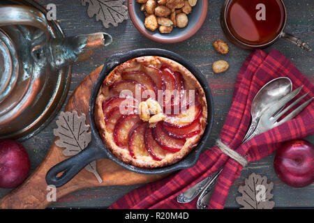 Tarte aux prunes maison cuit dans la poêle en fonte servi avec des arachides sur planche de bois. Vue de dessus avec de la vaisselle, de thé noir et de cuivre électrique sur le bois sombre. Banque D'Images