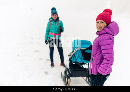 Maman avec poussette de bébé forêt profiter de l'hiver avec des ami ou partenaire, le temps de famille. La randonnée ou la marche sportive woman with sled la pram en bois. Banque D'Images