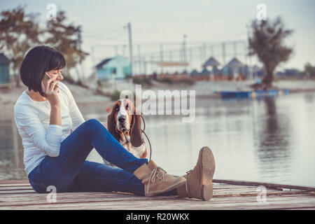 Portrait de belle jeune femme assise au bord de la rivière avec son chien Basset Hound tandis que talking on mobile phone Banque D'Images