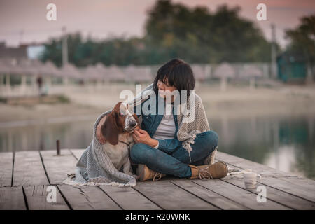 Portrait of beautiful young woman Playing with dog par la rivière. Femme heureuse avec son chien Basset Hound covered with blanket assis sur la jetée en bois Banque D'Images