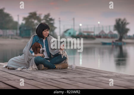 Portrait de belle jeune femme avec son chien recouvert d'une couverture, using digital tablet while sitting on wooden pier par la rivière. Coucher du soleil Banque D'Images