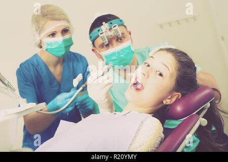 Portrait de peur teen girl avec la bouche ouverte pendant un traitement dentaire avec deux dentistes debout près de présidence Banque D'Images