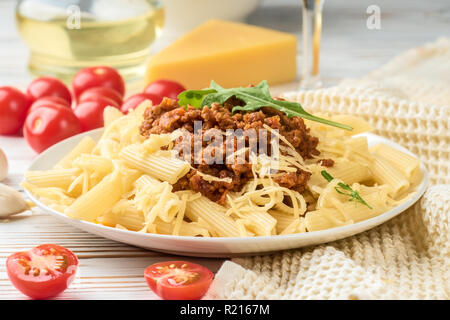 Les pâtes italiennes Penne bolognaise rigatone la viande hachée à la sauce tomate et de parmesan. Nature morte sur table en bois blanc, servi avec des tomates cerises, carafe d'huile d'olive et le verre de vin blanc Banque D'Images