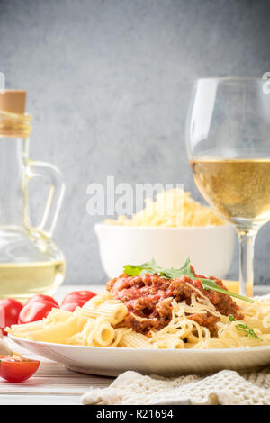 Les pâtes italiennes Penne bolognaise rigatone la viande hachée à la sauce tomate et de parmesan. Nature morte sur table en bois blanc, servi avec des tomates cerises, carafe d'huile d'olive et le verre de vin blanc Banque D'Images