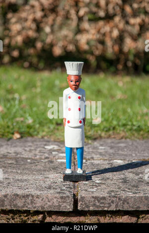 Petite figure en bois d'un chef teint foncé portant un manteau blanc et grand chapeau, debout sur des pavés en face d'une pelouse de gazon. Banque D'Images