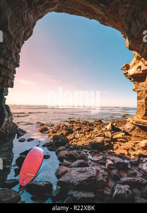 Abondoned Surf rouge à l'entrée d'une grotte marine. Beaucoup de roches au premier plan et un coucher de soleil paysage marin en arrière-plan. Situé le long de l'interface de coucher du soleil Banque D'Images