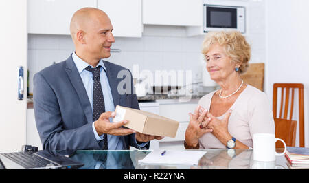 Représentant des ventes passant de carton avec des marchandises commandées à femme âgée dans la cuisine Banque D'Images