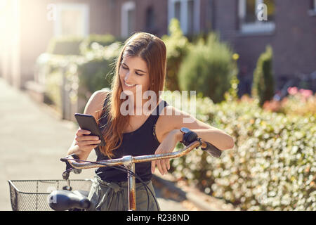 Belle jeune femme aux cheveux longs en demi-longueur/portrait en extérieur dans la ville debout appuyée sur vintage bicycle en utilisant smartphone moderne, l Banque D'Images