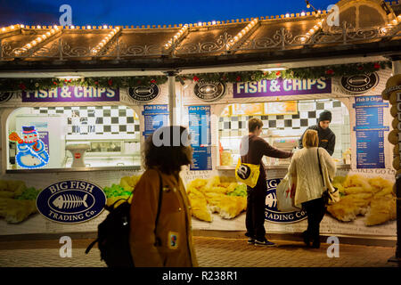 À un poisson-et-chip stall sur la jetée de Brighton un jeune homme et femme sont l'achat de nourriture. Le toit a lignes aériennes d'ampoules sous un ciel du soir. Banque D'Images