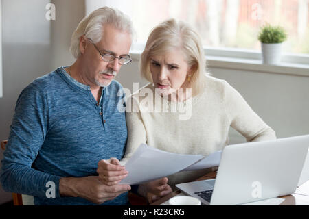 De graves âgés de mari et femme gérer les factures de services à l'aide d'ordinateur portable à la maison, couple concerné lire les documents hypothécaires prêt bancaire ou à une table de cuisine Banque D'Images