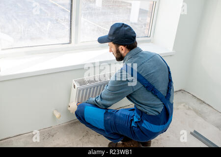 Montage du radiateur de chauffage de l'eau ouvrier près de la fenêtre dans le salon rénové blanc Banque D'Images