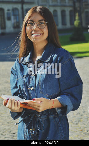 Un étudiant dans un costume en jean se tient sur le territoire de l'université et tient un livre ouvert dans ses mains, regarde smiling at the camera Banque D'Images