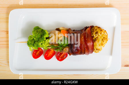 Vue de dessus de la caille grillée enveloppé dans jamon sur brochette servi sur plaque blanche avec des légumes et des verts sur fond de bois Banque D'Images