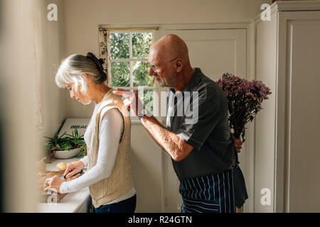 Vue latérale d'une femme travaillant dans la cuisine avec son mari debout derrière avec un bouquet de fleurs. Man se cache un bouquet de fleurs derrière lui pour Banque D'Images