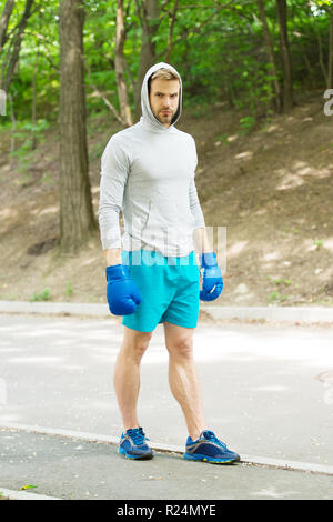 Grand jour arrive. Athlète homme concentré sur le visage avec des gants de boxe sport pratique prêt punch fond nature. Boxer hooded motivé pour réussir. Boxer sportif formation avec des gants de boxe. Banque D'Images