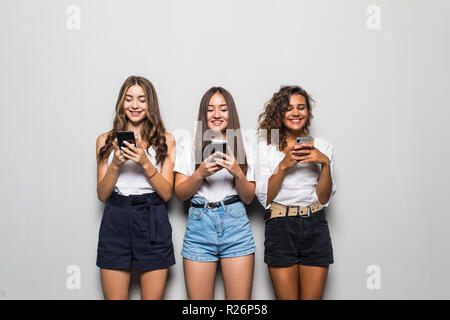 Trois belles jeunes filles portant des vêtements décontractés à la vers le haut et l'utilisation de téléphones cellulaires les plus isolées d'arrière-plan gris Banque D'Images